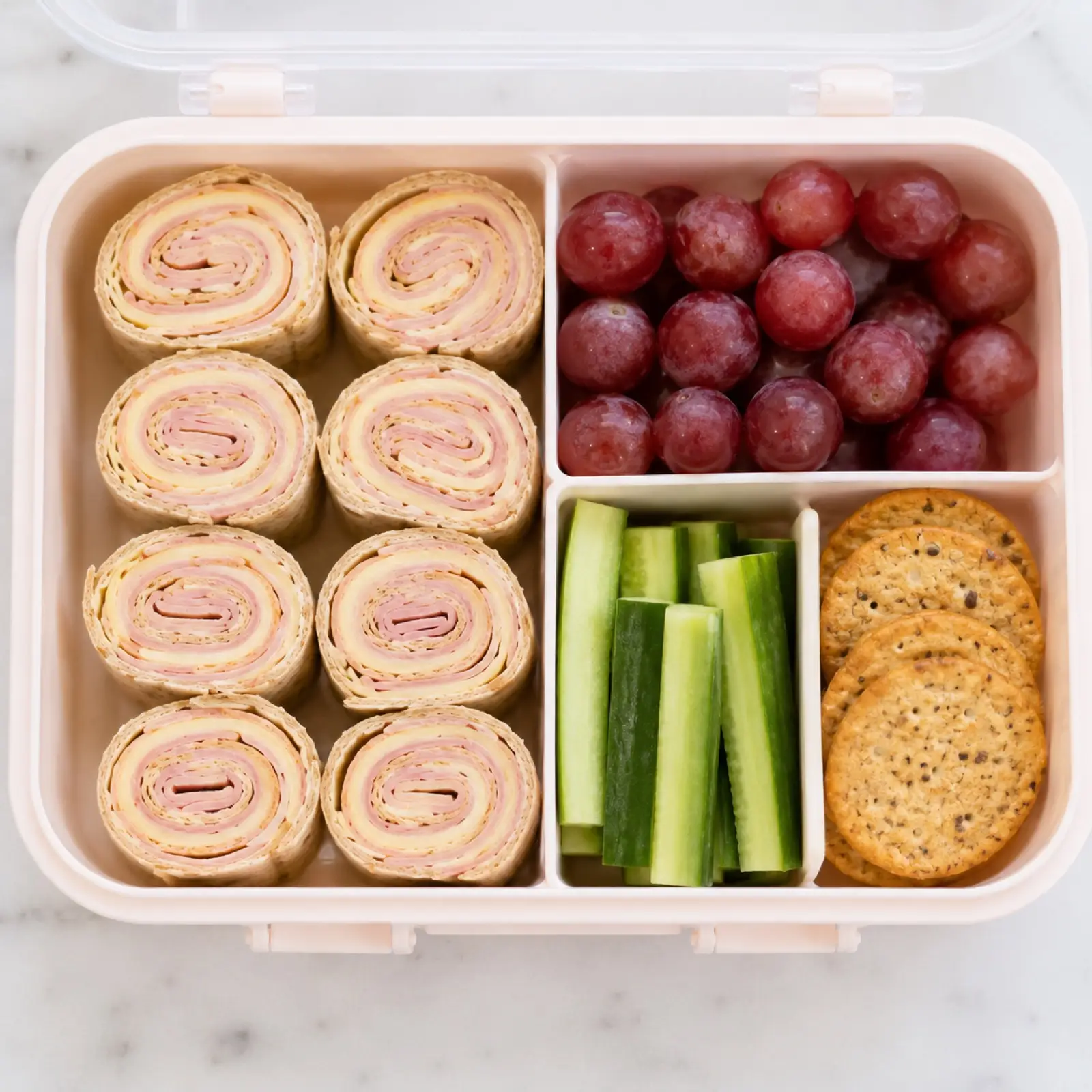 A kids bento lunchbox packed with whole wheat ham and cheese pinwheels, red grapes, cucumber sticks and whole grain crackers