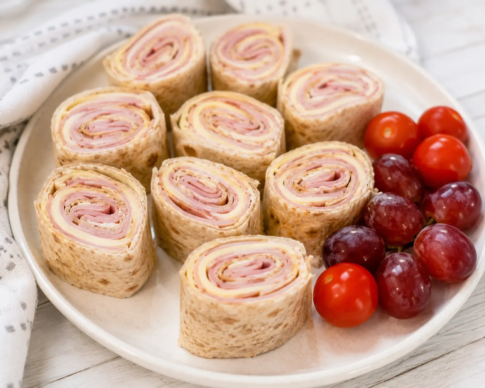 A plate of whole wheat tortilla ham and cheese roll-ups sliced into pinwheels showing the ham and cheese spiral, with cherry tomatoes and red grapes