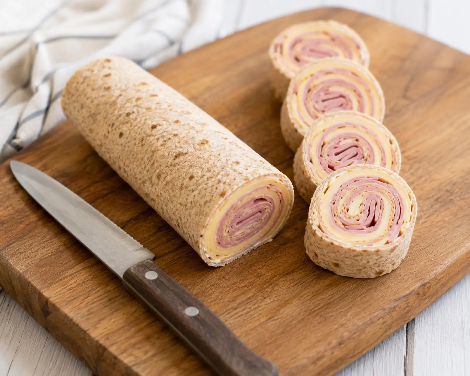 A rolled whole wheat tortilla with ham and cheese on a cutting board sliced into pinwheels showing the spiral cross-section, with a knife