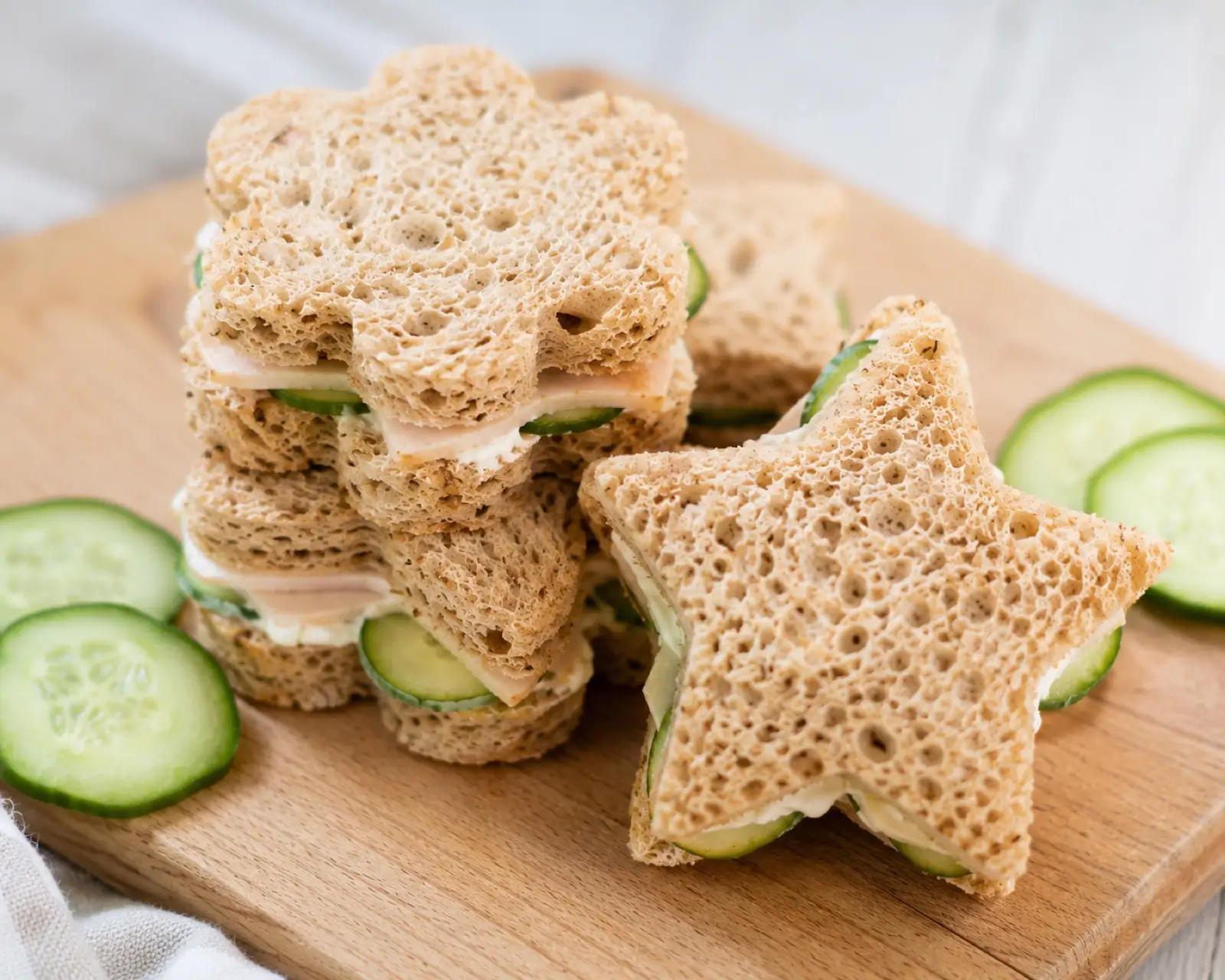 Close-up of star and flower shaped turkey cucumber cream cheese sandwiches on whole grain bread stacked on a wood board with cucumber slices
