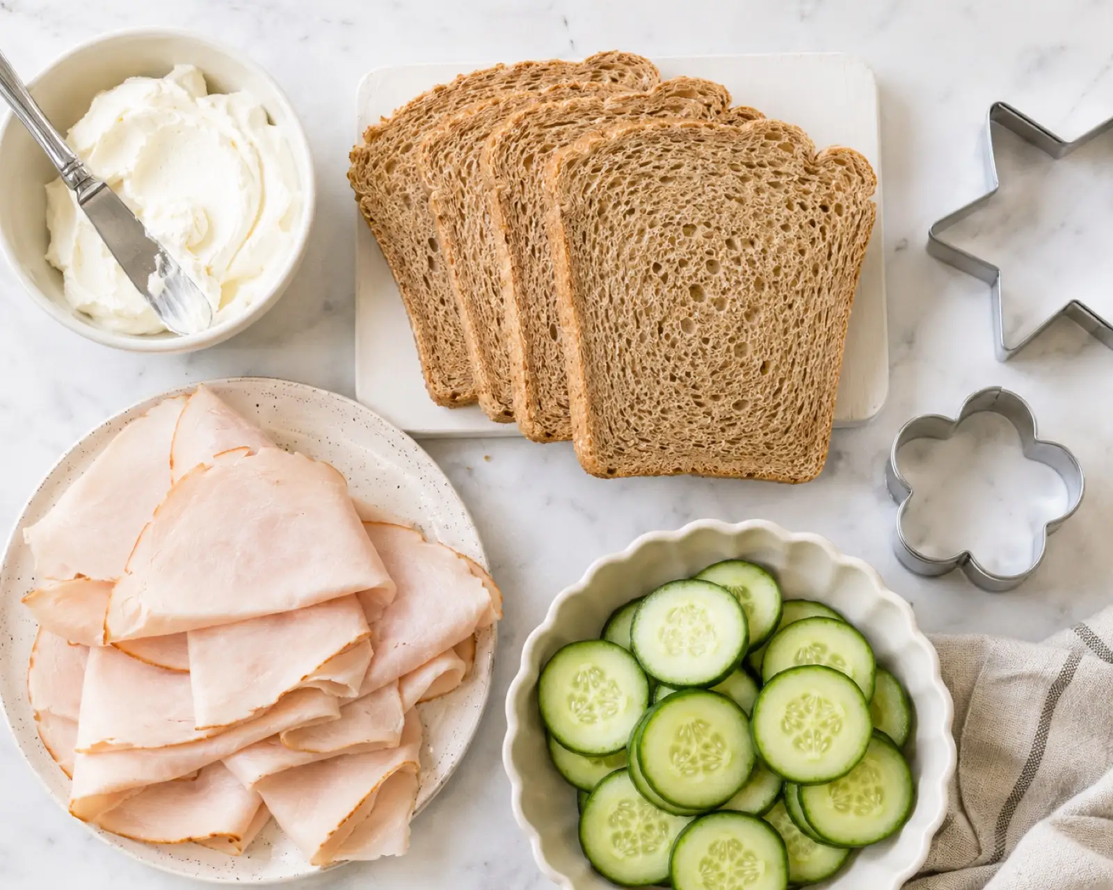Ingredients to make turkey cucumber cream cheese sandwiches: whole grain bread, cream cheese, sliced turkey, cucumber rounds and star and flower cookie cutters