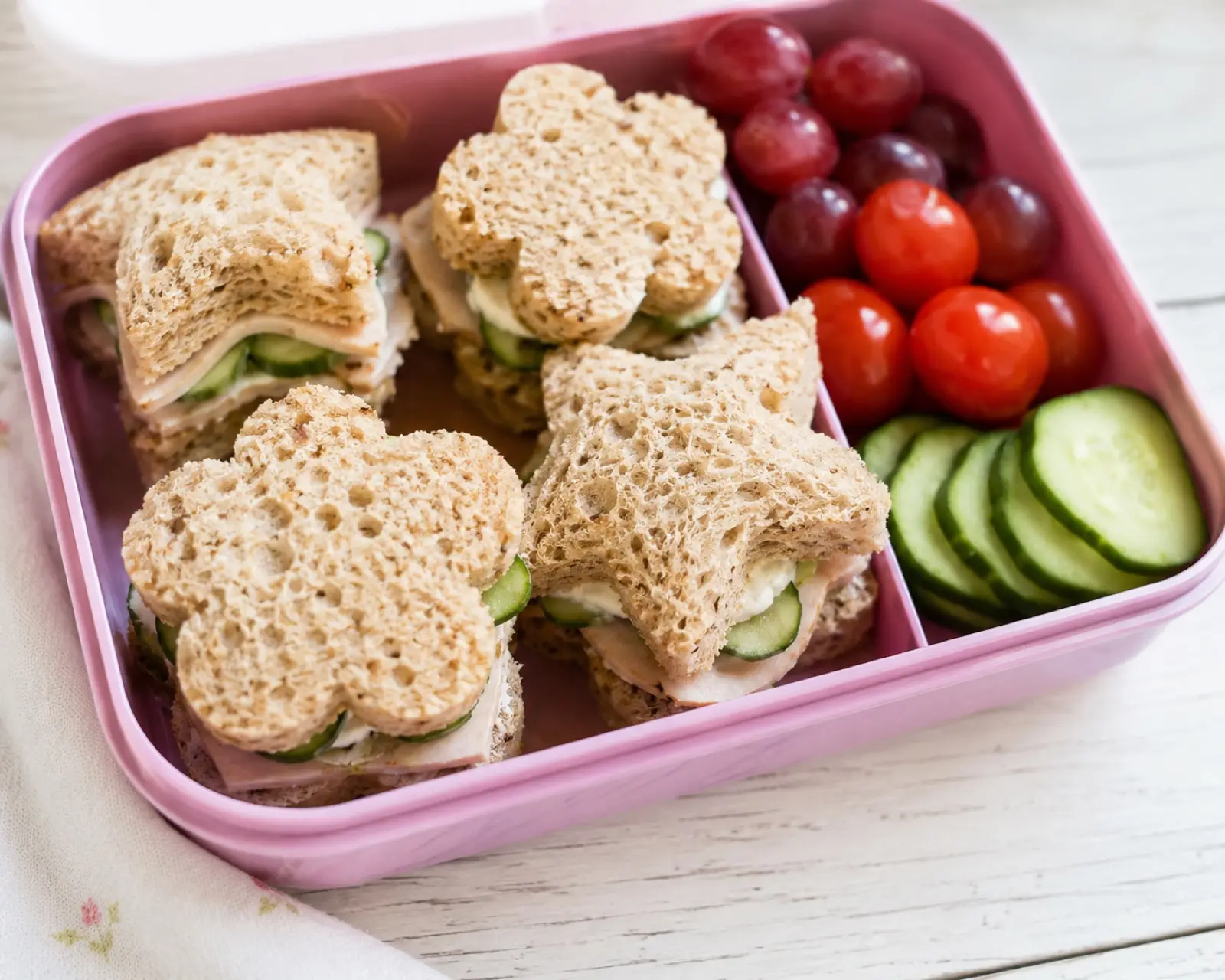 Bento lunchbox of turkey and cucumber cream cheese sandwiches on whole grain bread cut into star and flower shapes, with grapes, cherry tomatoes and cucumber slices